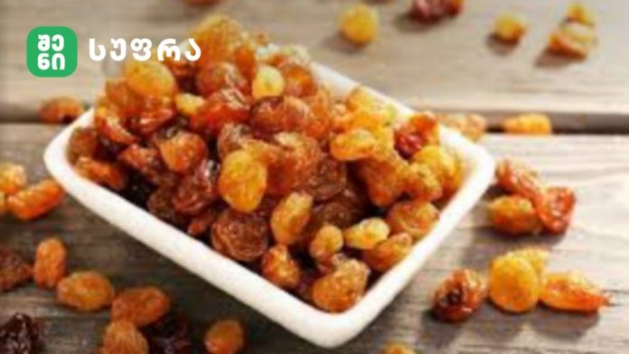 Tray of golden raisins on a wooden surface with a green logo in the top-left corner.