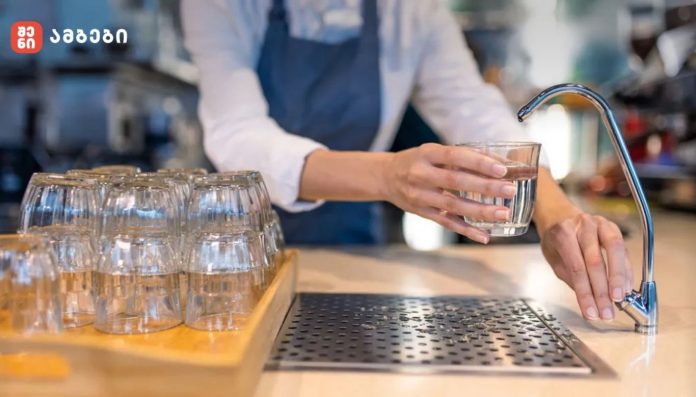 Person rinsing a glass under a kitchen faucet with a rack of glasses nearby on a wooden tray