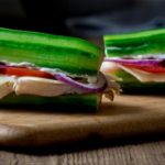 Two cucumber sandwiches filled with cheese, tomato, and red onion on a wooden cutting board.
