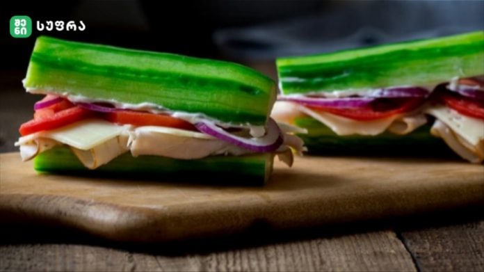 Two cucumber sandwiches filled with cheese, tomato, and red onion on a wooden cutting board.