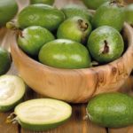Green feijoa fruits in a wooden bowl on a rustic wooden table, one cut open to show pale flesh inside.