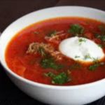Bowl of bright red beet soup (borscht) topped with sour cream and chopped dill; a garlic bulb in the background on the left.