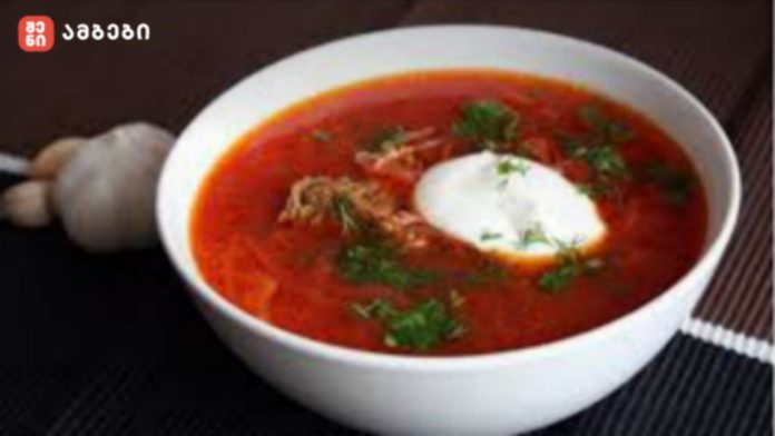 Bowl of bright red beet soup (borscht) topped with sour cream and chopped dill; a garlic bulb in the background on the left.