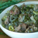 Bowl of beef stew with onions, green peppers and herbs in a light broth on a wooden table, with fresh greens beside it.