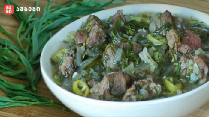 Bowl of beef stew with onions, green peppers and herbs in a light broth on a wooden table, with fresh greens beside it.