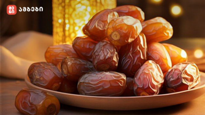 Plate of glossy brown dates piled on a shallow bowl, warm ambient lighting in the background, logo in the top-left corner.
