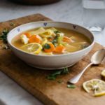 Bowl of lemon chicken soup with carrots and pasta on a wooden cutting board, with lemon wedges and a spoon nearby.