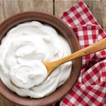 Bowl of creamy yogurt with a wooden spoon on a rustic wooden table, beside a red-and-white checkered napkin.