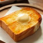 A slice of toasted bread with a pat of butter on a white napkin, placed on a wooden plate, ready for breakfast.
