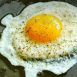 Sunny-side-up fried egg cooking in a skillet with herbs and oil around the edges.