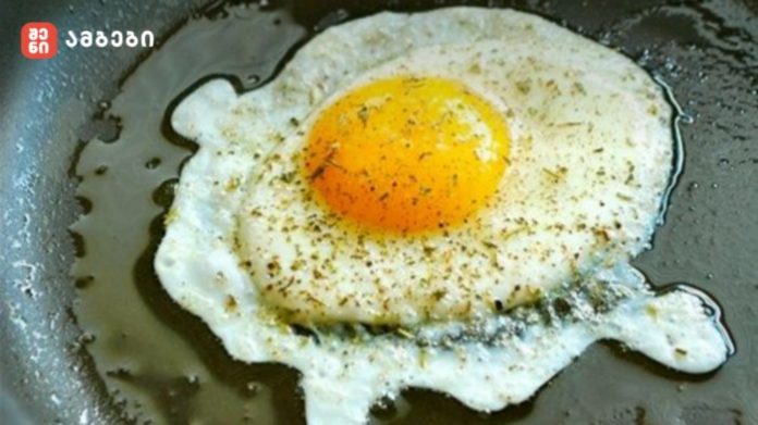 Sunny-side-up fried egg cooking in a skillet with herbs and oil around the edges.