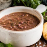 White ceramic bowl of hearty bean soup with a toasted bread slice on a wooden board, garnished with parsley.