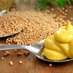 Spoon with swirls of yellow mustard on a metal spoon, with mustard seeds scattered on a wooden surface nearby.