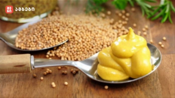 Spoon with swirls of yellow mustard on a metal spoon, with mustard seeds scattered on a wooden surface nearby.