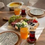 A wooden dining table set for breakfast with a cheese, grape, and meat platter, orange and red drinks, a teapot, and stacked plates nearby.