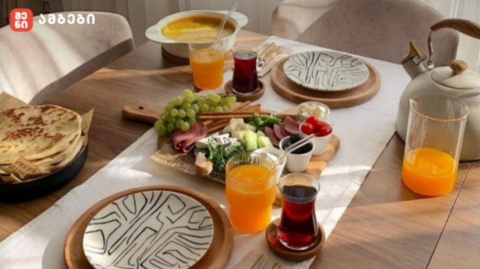 A wooden dining table set for breakfast with a cheese, grape, and meat platter, orange and red drinks, a teapot, and stacked plates nearby.