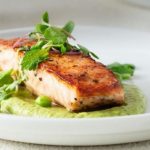 Pan-seared salmon fillet on a green pea-mint sauce with peas and microgreens on a white plate.
