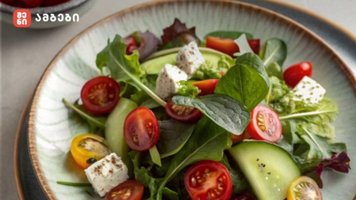Fresh mixed green salad with cherry tomatoes, cucumber slices, and feta cheese on a plate; logo in the top-left corner.