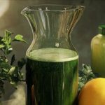 Green juice in a glass pitcher surrounded by fresh herbs and citrus on a kitchen counter