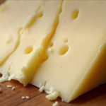Wedge of yellow cheese sliced on a wooden board, with small cheese crumbles around it.