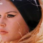 Close-up of a blonde woman with winged eyeliner, wearing a black headband, outdoors in sunlight.