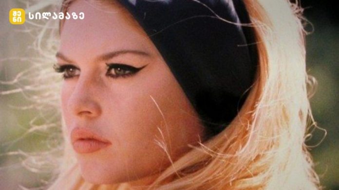 Close-up of a blonde woman with winged eyeliner, wearing a black headband, outdoors in sunlight.