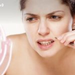 Woman plucking eyebrow hair with tweezers while looking in a round pink mirror to her left.