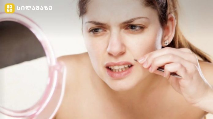Woman plucking eyebrow hair with tweezers while looking in a round pink mirror to her left.