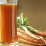 Glass of orange carrot juice on a wooden surface with fresh carrots in the background, suggesting a fresh carrot juice setup
