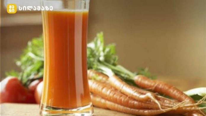 Glass of orange carrot juice on a wooden surface with fresh carrots in the background, suggesting a fresh carrot juice setup