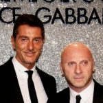 Two men in black tuxedos pose for a photo at a Dolce & Gabbana event, standing before a glittery backdrop.