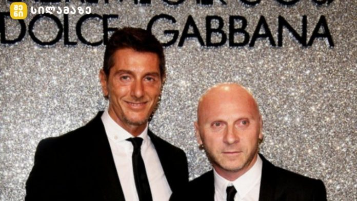 Two men in black tuxedos pose for a photo at a Dolce & Gabbana event, standing before a glittery backdrop.