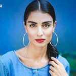 Portrait of a woman with dark hair in a ponytail, wearing large hoop earrings and a blue denim top against a blue background.