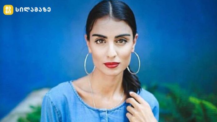 Portrait of a woman with dark hair in a ponytail, wearing large hoop earrings and a blue denim top against a blue background.