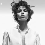 Black-and-white fashion portrait of an androgynous model in an open white shirt, looking straight ahead; logo top-left corner.