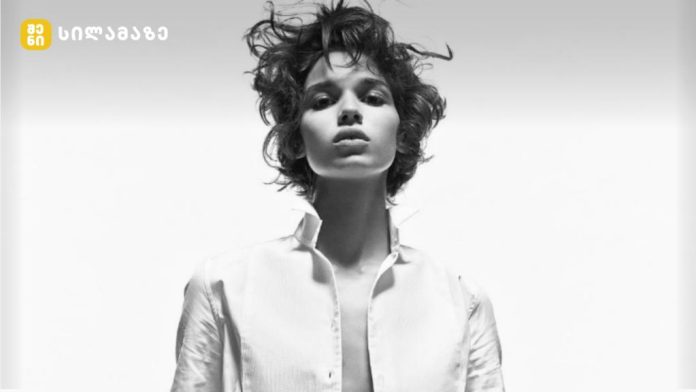Black-and-white fashion portrait of an androgynous model in an open white shirt, looking straight ahead; logo top-left corner.