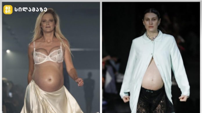 Two pregnant models walk a fashion show runway, one in lace bra and satin skirt, the other in an open shirt revealing a baby bump.