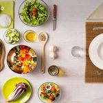 Top-down view of a colorful kitchen spread with tomatoes, cucumbers, peppers, bowls of chopped veggies, and a person setting a plate at the table