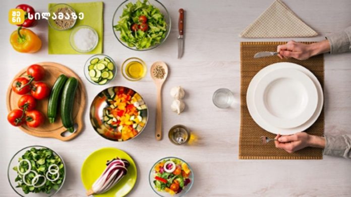 Top-down view of a colorful kitchen spread with tomatoes, cucumbers, peppers, bowls of chopped veggies, and a person setting a plate at the table