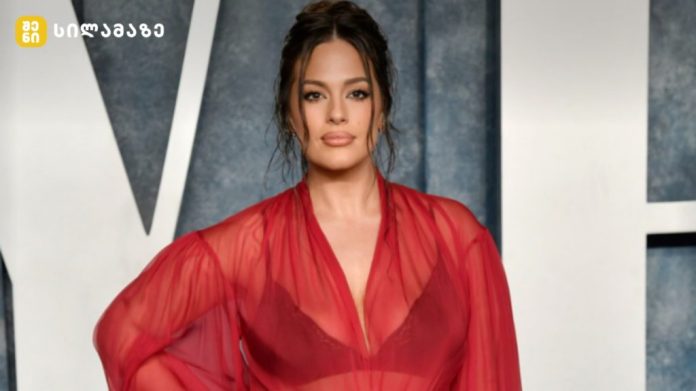 Woman wearing a sheer red dress posing on a red carpet at a formal event, dark hair pulled back and subtle makeup.