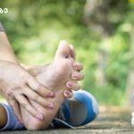 Person sits outdoors and massages their foot, with blue sneakers and a water bottle nearby on a park path.