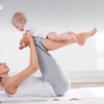 Mother lying on a white mat while lifting her baby overhead with legs raised in a calm, sunlit room.