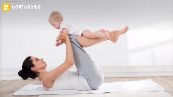 Mother lying on a white mat while lifting her baby overhead with legs raised in a calm, sunlit room.