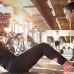 Two women workout in a gym: one lying on a mat doing sit-ups while the trainer crouches beside her offering support, with weights in the background.