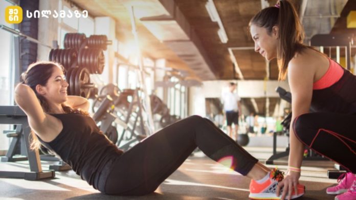 Two women workout in a gym: one lying on a mat doing sit-ups while the trainer crouches beside her offering support, with weights in the background.