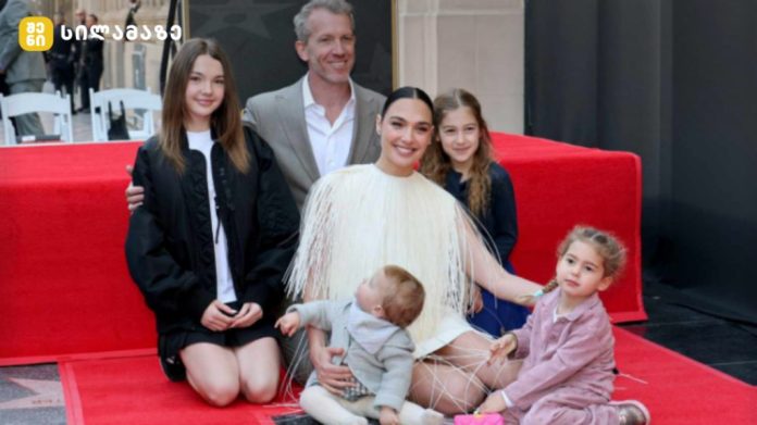 A woman in a white fringe dress sits on a red carpet surrounded by children and adults, posing for a photo at an event.