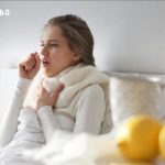Woman in bed coughing into her elbow, wrapped in a scarf, with a blanket close by and bedside items like lemons and a teapot nearby.