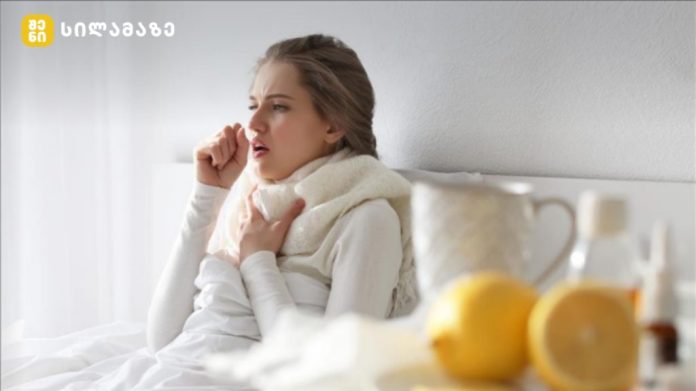 Woman in bed coughing into her elbow, wrapped in a scarf, with a blanket close by and bedside items like lemons and a teapot nearby.
