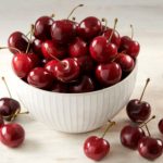 White ceramic bowl brimming with ripe red cherries on a light wooden surface disabling the cluttered background, fresh and glossy.