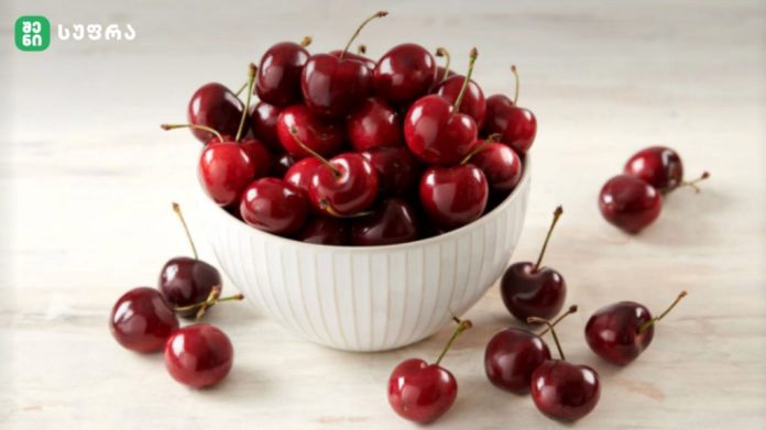 White ceramic bowl brimming with ripe red cherries on a light wooden surface disabling the cluttered background, fresh and glossy.